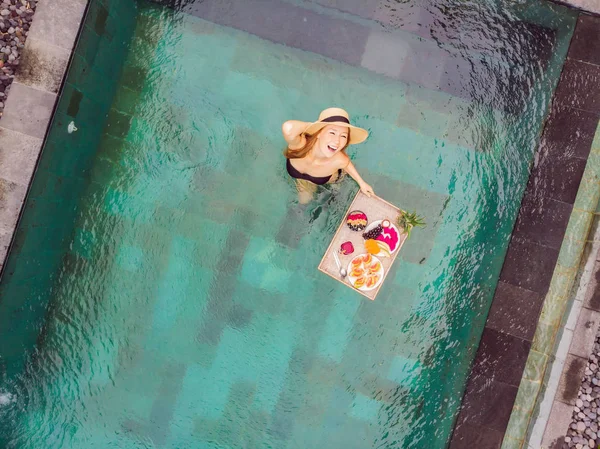 Breakfast tray in swimming pool, floating breakfast in luxury hotel. Girl relaxing in the pool drinking smoothies and eating fruit plate, smoothie bowl by the hotel pool. Exotic summer diet. Tropical