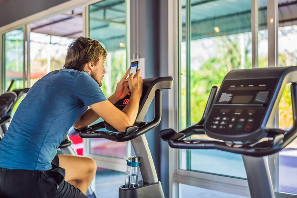 Lifestyle portrait of handsome man with phone sitting on the simulator in the gym — Stock Photo, Image
