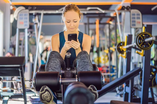 Молодая женщина пользуется телефоном во время тренировки в спортзале. Женщина сидит на тренажере с мобильным телефоном — стоковое фото