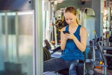 Spor salonunda eğitim görürken telefonu kullanan genç bir kadın. Egzersiz makinesinde oturan bir kadın cep telefonu tutuyor.