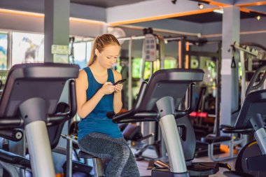 Spor salonunda eğitim görürken telefonu kullanan genç bir kadın. Egzersiz makinesinde oturan bir kadın cep telefonu tutuyor.