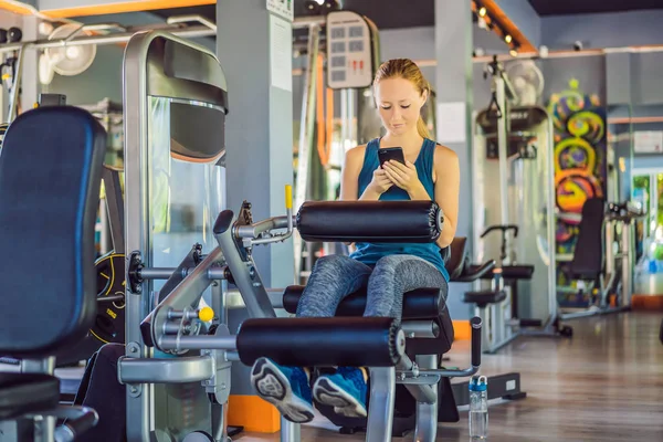 Молодая женщина пользуется телефоном во время тренировки в спортзале. Женщина сидит на тренажере с мобильным телефоном — стоковое фото