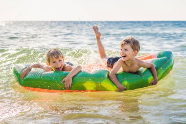Çocuklar şişme bir şilte üzerinde denizde yüzer ve eğlenirler.