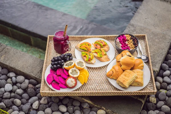 Café da manhã em uma bandeja com frutas, pães, sanduíches de abacate, tigela de smoothie na piscina. Dieta saudável de verão, café da manhã vegan. Conceito de férias saborosas — Fotografia de Stock