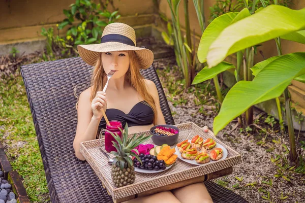 Young woman eating breakfast in a lounge chair on a tray with fruit, buns, avocado sandwiches, smoothie bowl by the pool. Summer healthy diet, vegan breakfast. Tasty vacation concept