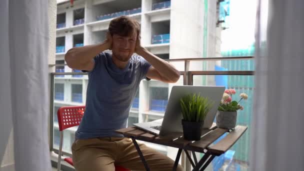 Slowmotion shot van een jonge man zittend op een balkon met een notebook en lijden aan een luide ruis geproduceerd door een bouwplaats in de buurt. Concept van geluidsoverlast in grote steden — Stockvideo