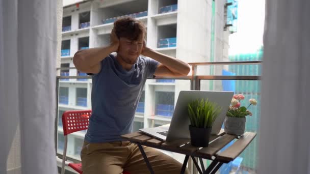 Foto lenta de um jovem sentado em uma varanda com um caderno e sofrendo de um barulho alto produzido por um canteiro de obras nas proximidades. Conceito de poluição sonora nas grandes cidades — Vídeo de Stock