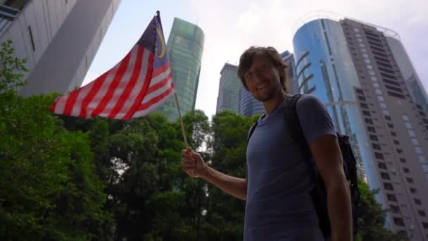 Fotografía en cámara lenta de un joven que sostiene un teléfono inteligente y ondea la bandera de Malasia con rascacielos en un segundo plano. Viajar a Malasia concepto — Vídeos de Stock