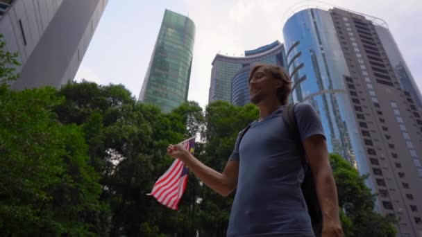 Fotografía en cámara lenta de un joven que sostiene un teléfono inteligente y ondea la bandera de Malasia con rascacielos en un segundo plano. Viajar a Malasia concepto — Vídeos de Stock