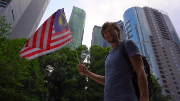 Fotografía en cámara lenta de un joven que sostiene un teléfono inteligente y ondea la bandera de Malasia con rascacielos en un segundo plano. Viajar a Malasia concepto — Vídeos de Stock