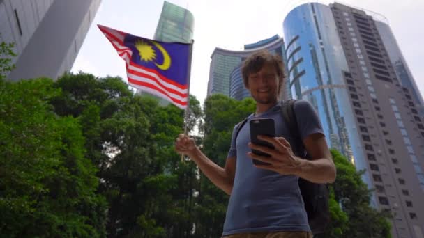 Slowmotion shot van een jonge man die een smartphone en golven Maleisische vlag met wolkenkrabbers op een achtergrond houdt. Reizen naar Maleisië concept — Stockvideo