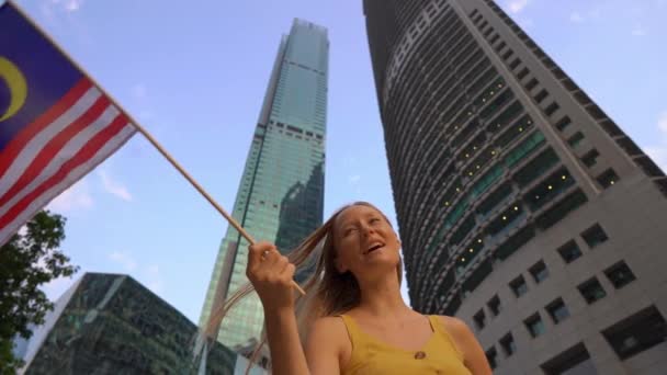 Fotografía en cámara lenta de una joven que ondea la bandera de Malasia con rascacielos en un segundo plano. Viajar a Malasia concepto — Vídeos de Stock