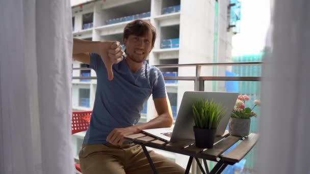 Slowmotion shot van een jonge man zittend op een balkon met een notebook en lijden aan een luide ruis geproduceerd door een bouwplaats in de buurt. Concept van geluidsoverlast in grote steden — Stockvideo