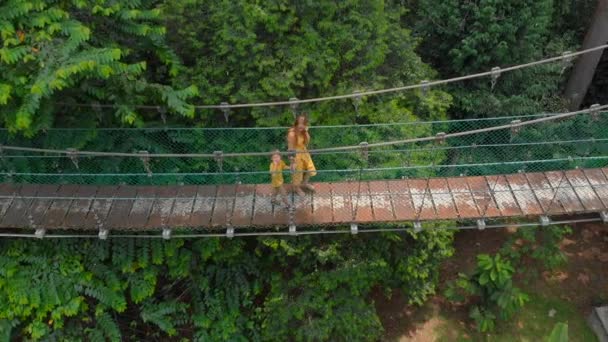 Un disparo aéreo de una mujer joven y su hijo pequeño caminando sobre un puente de suspensión sobre las selvas. Vacaciones en el concepto tropical — Vídeo de stock