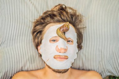 Genç adam salyangoz sümüğünden yüz maskesi yapıyor. Maskenin üzerinde sürünen bir salyangoz. Erkekler için spa, herkes için spa