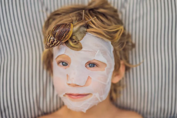 Fortement plus jeune du masque avec du mucus d'escargot. Un enfant dans un masque pour le visage avec un escargot. Escargot rampant sur un masque facial. SPA pour tous — Photo