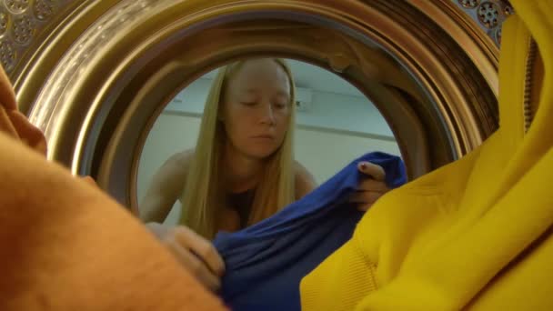 Utsikt från tvätt maskinen som en ung kvinna sätter smutsiga kläder i det — Stockvideo