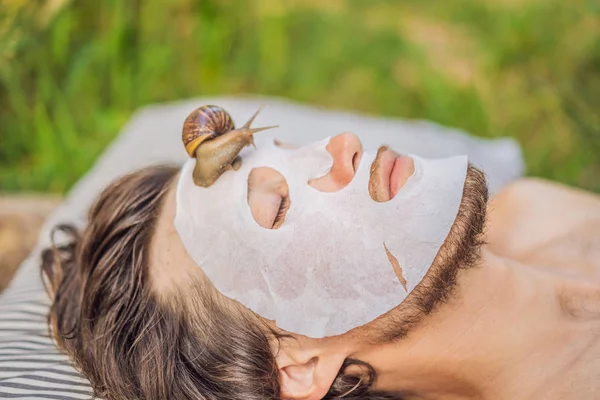 Genç adam salyangoz sümüğünden yüz maskesi yapıyor. Maskenin üzerinde sürünen bir salyangoz. Erkekler için spa, herkes için spa — Stok fotoğraf