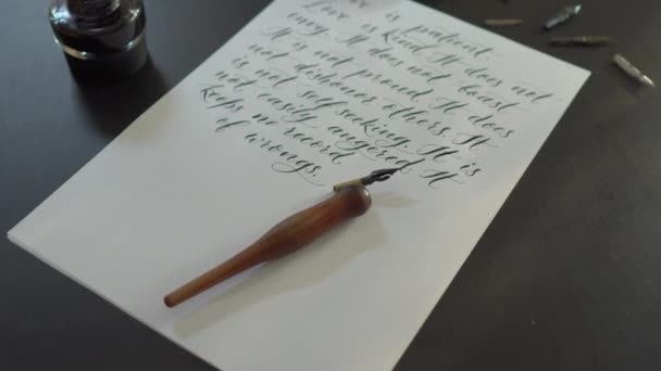 Tiro de close-up de uma caligrafia ferramentas e uma garrafa de tinta que coloca em torno de uma folha de um livro branco com várias linhas de uma Bíblia sobre o amor está escrito nele — Vídeo de Stock