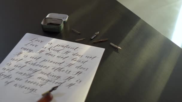 Closeup shot of a calligraphy tools and a bottle of ink laying around a sheet of a white paper with several lines from a Bible about love is written on it — Stock Video