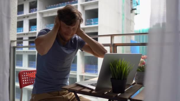 Young man sitting on a balcony with a notebook and suffering from a loud noise produced by a construction site nearby. Concept of noise pollution in big cities — Stock Video