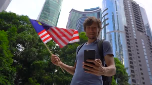 Um jovem segura um smartphone e acena bandeira da Malásia com arranha-céus em um fundo. Viagem ao conceito da Malásia — Vídeo de Stock