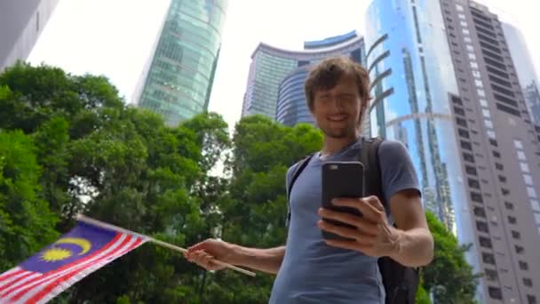 En ung mann har en smarttelefon og bølger med malaysisk flagg med skyskrapere i bakgrunnen. Reise til Malaysia-konseptet – stockvideo