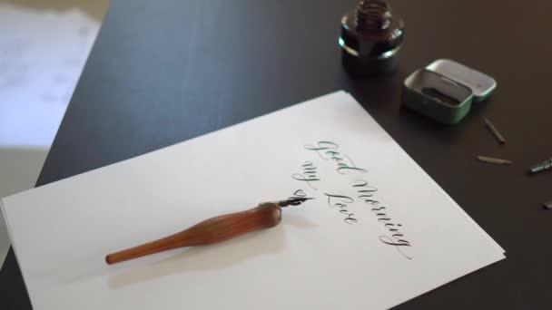 Close-up shot van een kalligrafiegereedschap en een fles inkt leggen rond een vel van een Witboek met Goedemorgen mijn liefde geschreven op het — Stockvideo