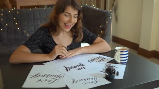 Крупный план молодой женщины-каллиграфа, пишущей на бумаге с использованием техники письма. Она смотрит на листы со словами "Люби себя" и смеется — стоковое видео