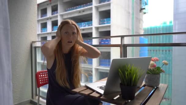 Slowmotion shot of a young woman sitting on a balcony with a notebook and suffering from a loud noise produced by a construction site nearby. Concept of noise pollution in big cities — Stock Video