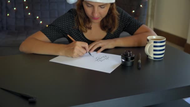 Nahaufnahme einer jungen Frau mit Weihnachtsmütze, die in Schrifttechnik auf ein Papier schreibt. Sie schreibt frohe Weihnachten — Stockvideo