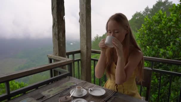 Slowmotion tiro de uma jovem mulher bebendo chá ou café sentado em um café nas montanhas com uma vista sobre uma montanha Batur e sua caldeira escondida nas nuvens. Conceito de Travell para Bali — Vídeo de Stock