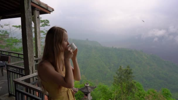 Zeitlupenaufnahme einer jungen Frau, die in einem Café in den Bergen Tee oder Kaffee trinkt, mit Blick auf eine Bergbatur und ihre in Wolken versteckte Caldera. travell to bali concept — Stockvideo