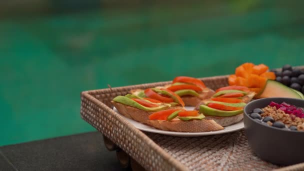 Tiro de close-up de um café da manhã pessoal em uma mesa flutuante em um lado de uma piscina privada. Um menino entra em cena e tira um batido de frutas da mesa — Vídeo de Stock