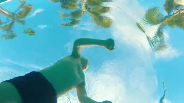 View from a bottom of a swimming pool on palm trees and a small kid swimming over the camera — Stock Video