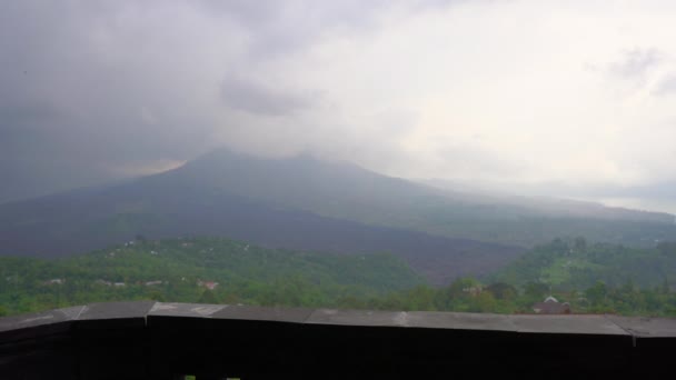 Una giovane donna in abito giallo e cappello visita un belvedere con vista su una montagna Batur e la sua caldera nascosta tra le nuvole. Viaggiare a Bali concetto — Video Stock