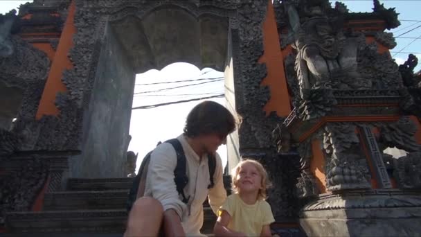 Slowmotion shot of a father and son tourists sitting on the stairs of a Balinese temple making a selfie on an action camera — Stock Video