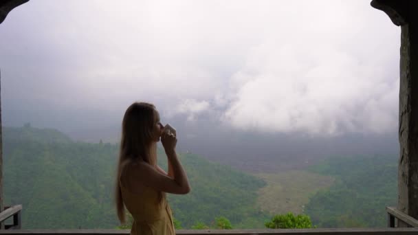 Fotografía en cámara lenta de una joven bebiendo té o café en un café en las montañas con vistas a una montaña Batur y su caldera escondida en las nubes. Viajar al concepto de Bali — Vídeos de Stock