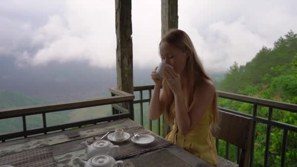 Fotografía en cámara lenta de una joven bebiendo té o café sentada en un café en las montañas con vistas a una montaña Batur y su caldera escondida en las nubes. Viajar al concepto de Bali — Vídeo de stock