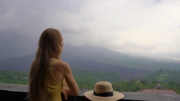 Sarı elbiseli ve şapkalı genç bir kadın, batur dağına ve bulutlara gizlenmiş kalderasına bir bakış açısıyla bakar. Travell bali kavramı — Stok video