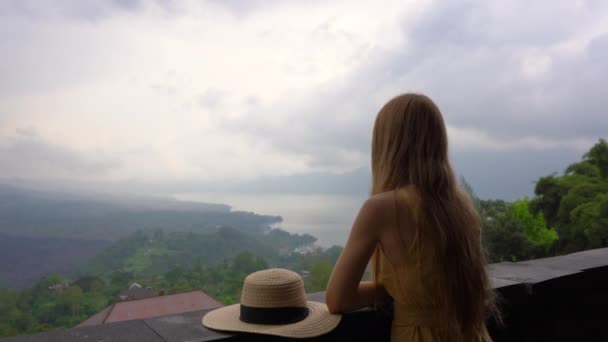 Una giovane donna in abito giallo e cappello visita un belvedere con vista su una montagna Batur e la sua caldera nascosta tra le nuvole. Viaggiare a Bali concetto — Video Stock