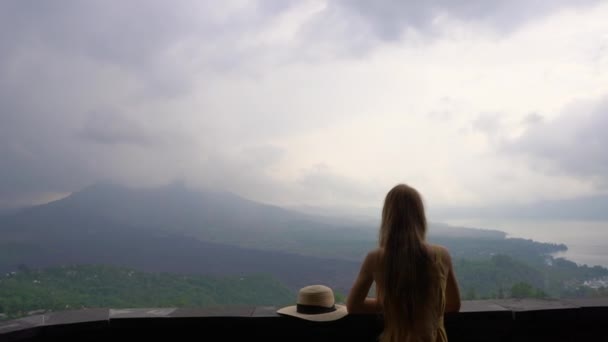 一位身着黄色连衣裙、头戴帽子的年轻女子，在云中欣赏巴图尔山及其火山口的景色。前往巴厘岛概念 — 图库视频影像
