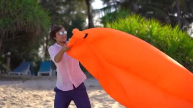 Tropik bir sahilde genç adam şişme kanepe şişirir. Yaz tatili konsepti