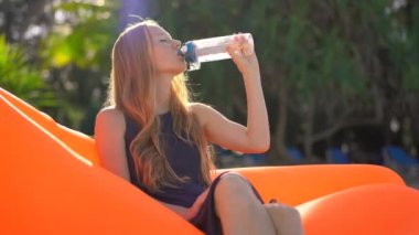 Tropik bir plajda genç kadın şişme bir kanepe üzerinde oturur ve çok kullanılabilir plastik şişe su içecekler. Biraz daha su iç. Yeniden kullanılabilir plastik ve plastik atık kavramını azaltın
