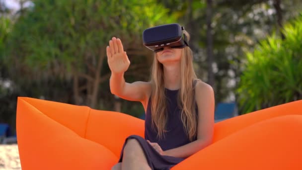 Junge Frau sitzt auf einem aufblasbaren Sofa an einem tropischen Strand und trägt eine Vr-Brille. Konzept moderner Technologien, die Ihnen das Gefühl geben können, sich woanders zu befinden — Stockvideo