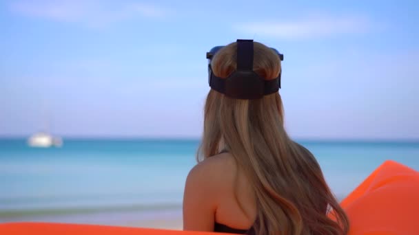 Junge Frau sitzt auf einem aufblasbaren Sofa an einem tropischen Strand und trägt eine Vr-Brille. Konzept moderner Technologien, die Ihnen das Gefühl geben können, sich woanders zu befinden — Stockvideo
