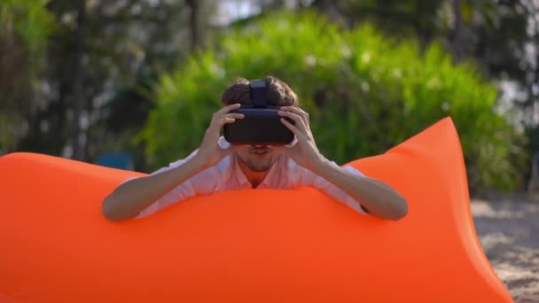 Jovem em um sofá inflável em uma praia tropical usa um óculos VR. Ele se sente como se estivesse nadando em um mar observando muitos peixes tropicais. Conceito de tecnologias modernas que podem fazer você se sentir — Vídeo de Stock