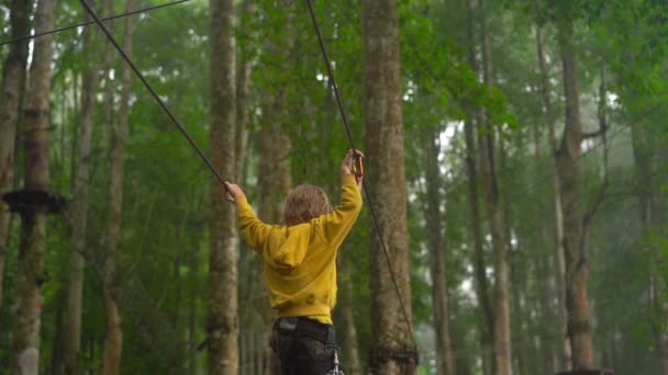 Superslowmotion skott av en liten pojke i en säkerhetssele klättrar på en rutt i trädtopparna i en skog äventyrspark. Han klättrar på hög repspår. Nöjescentrum utomhus med klättrings aktiviteter — Stockvideo