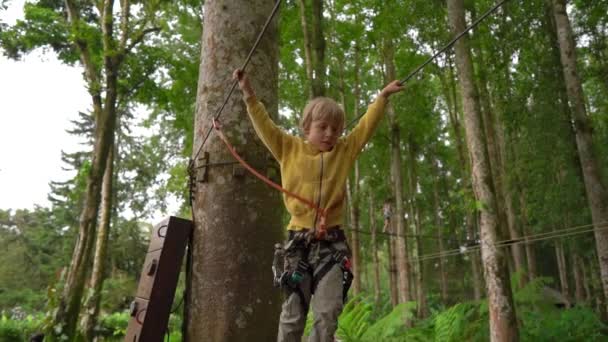 在森林探险公园的树梢上，一个小男孩在安全带上攀爬的超慢动作镜头。他爬上高高的绳索。户外娱乐中心，有登山活动 — 图库视频影像