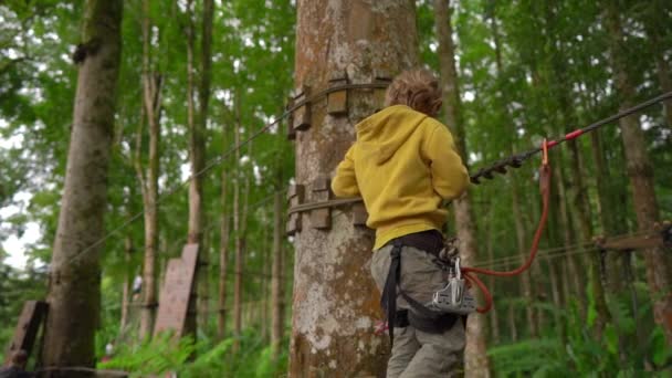 Superslowmotion plan d'un petit garçon dans un harnais de sécurité grimpe sur un chemin dans la cime des arbres dans un parc d'aventure forestière. Il grimpe sur le sentier des hautes cordes. Centre de loisirs extérieur avec activités d'escalade — Video